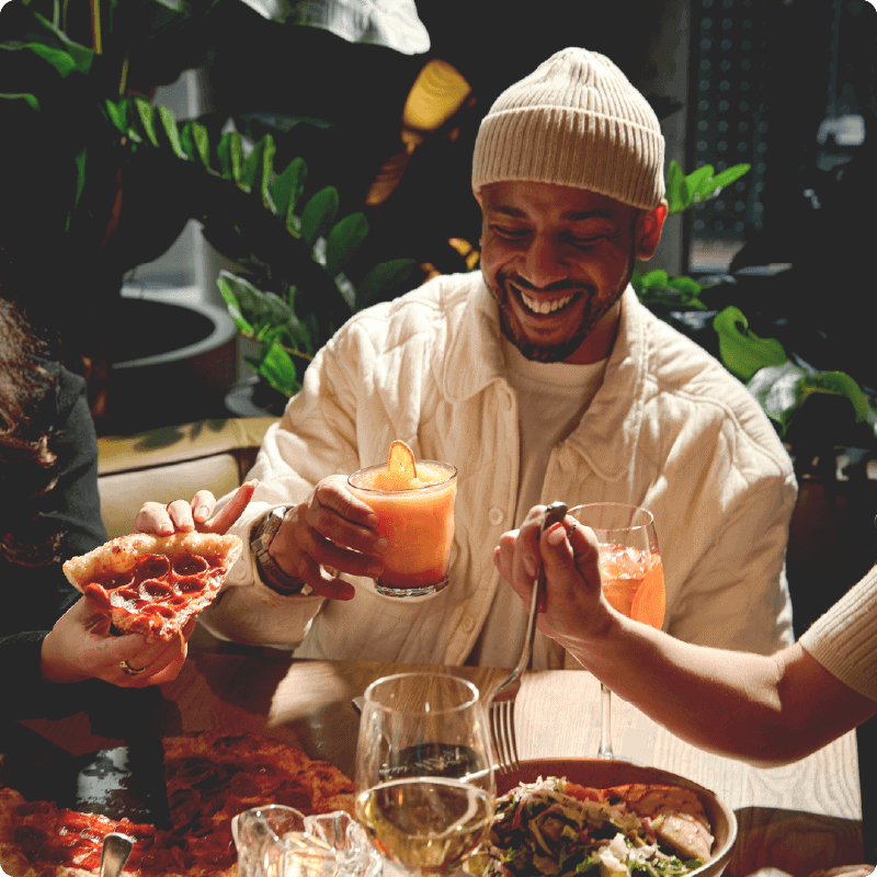Person holding a cocktail over a table of pizza with friends